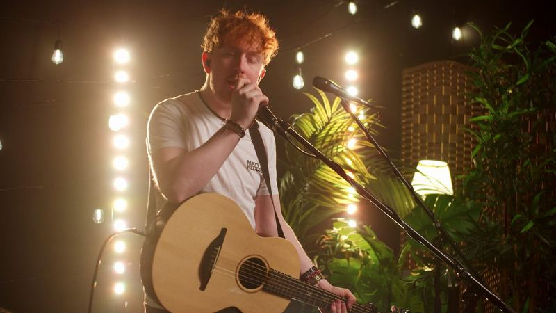 Matt Keddie singing into the mic with an acoustic guitar under warm festoon lights